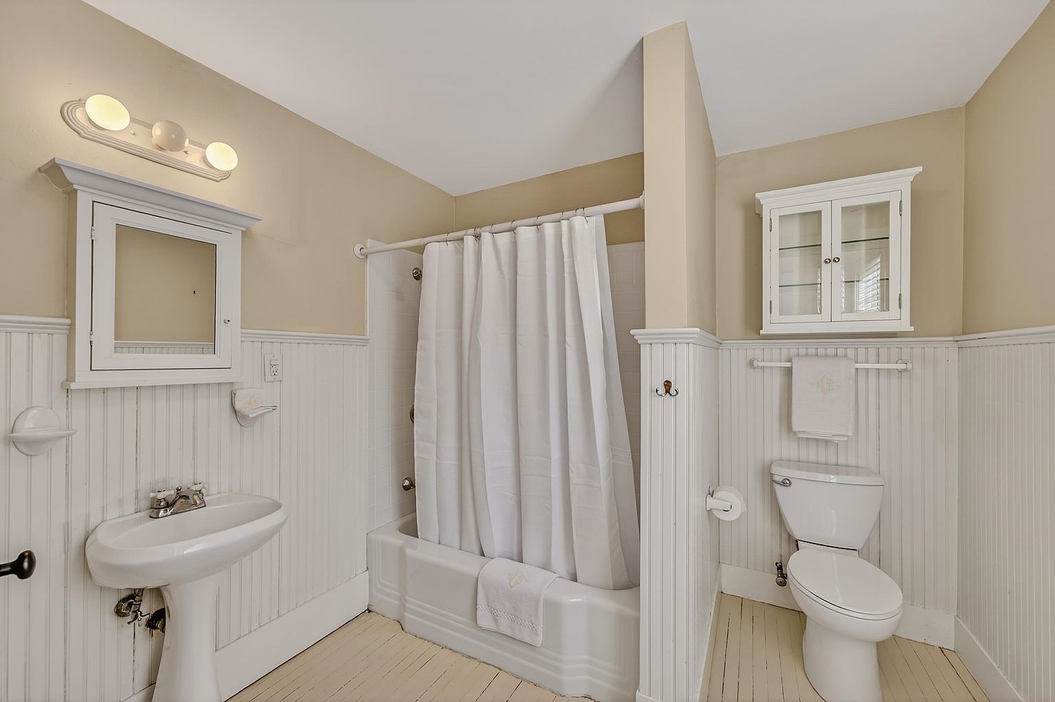 This bright, vintage-inspired bathroom features a classic pedestal sink, a white tub-shower combination with a curtain, and a standard toilet. The space is accented with white beadboard wainscoting, a wall-mounted medicine cabinet, and a small glass-front wall cabinet for storage. The room is finished with light-toned wood flooring and a simple three-light vanity fixture, creating a clean and functional aesthetic.