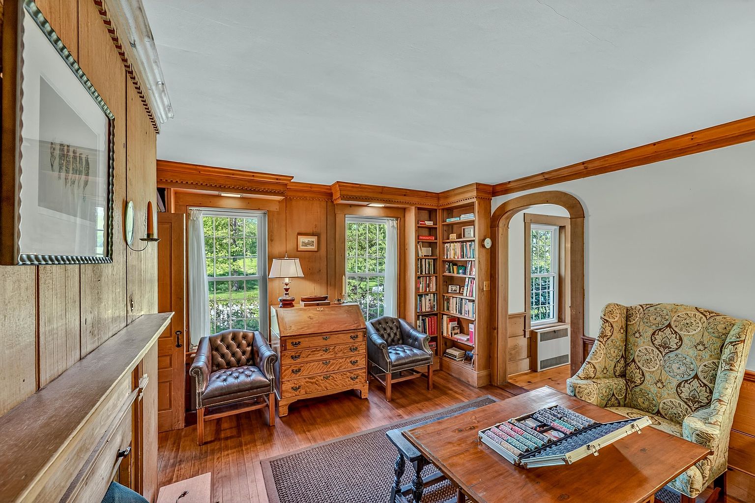 This cozy, traditional study features rich wood-paneled walls, built-in bookshelves, and warm hardwood flooring. The room is furnished with two leather armchairs, a classic secretary desk, and a patterned armchair, creating a sophisticated and scholarly atmosphere. Large windows allow natural light to fill the space, highlighting the intricate woodwork and the inviting, library-like aesthetic.
