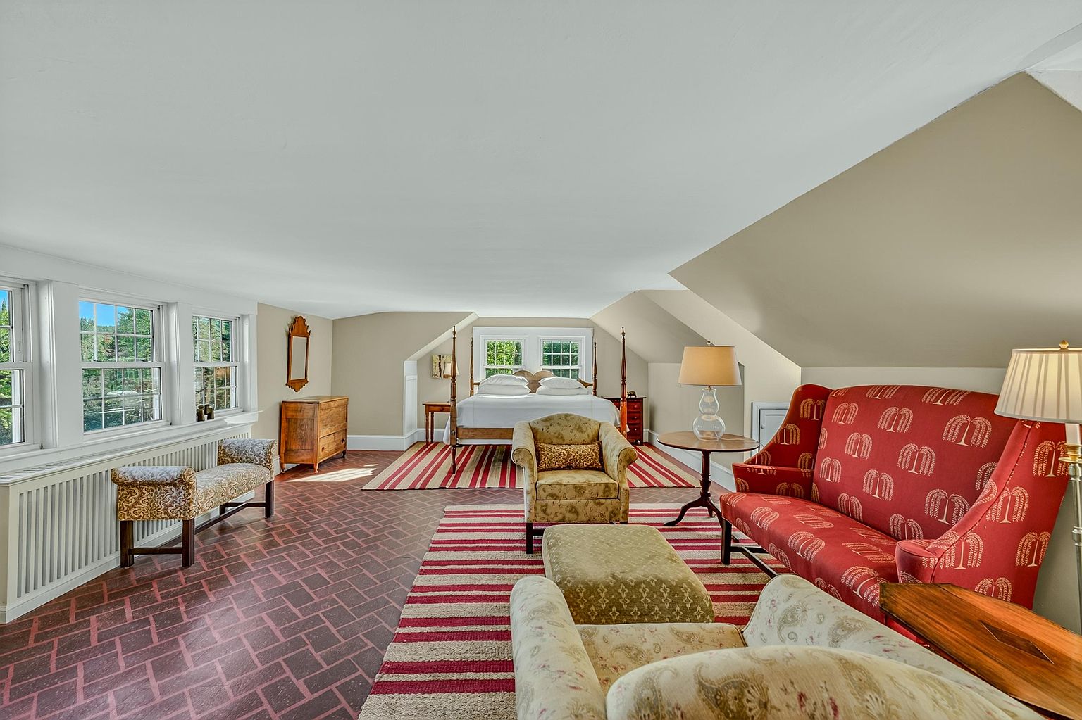 This expansive primary bedroom features a unique layout with a brick-patterned floor and a vaulted ceiling, creating a spacious and airy atmosphere. The room is furnished with a four-poster bed, a vibrant red patterned sofa, and multiple seating areas, all anchored by striped area rugs. Large windows along the side wall provide ample natural light, while the neutral wall tones offer a balanced backdrop for the eclectic furniture arrangement.