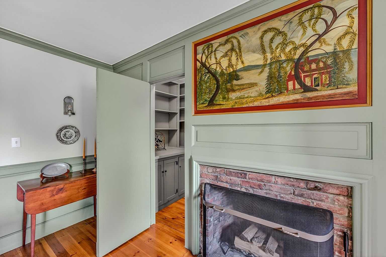 This interior view showcases a cozy living space featuring a brick fireplace with a black metal screen and a large, framed landscape painting mounted above it. The walls are painted a soft sage green, complemented by warm-toned wood flooring and a wooden console table adorned with decorative plates. A partially open door reveals a glimpse into a secondary room with built-in shelving and gray cabinetry, creating a sense of depth and architectural interest.