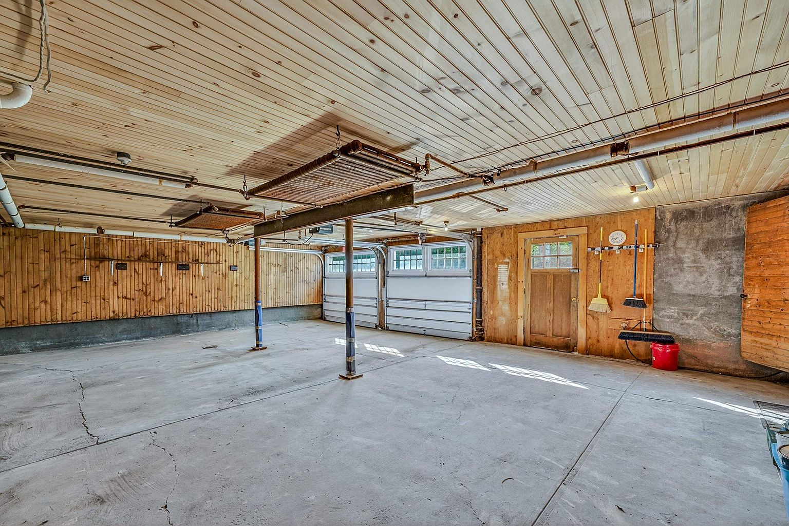 This spacious, multi-car garage features a concrete floor and walls lined with light-toned wood paneling. The ceiling is finished with matching wood planks, and the space includes two garage doors, a wooden entry door, and exposed structural support beams. The overall aesthetic is rustic and functional, providing ample room for vehicle storage or a workshop area.
