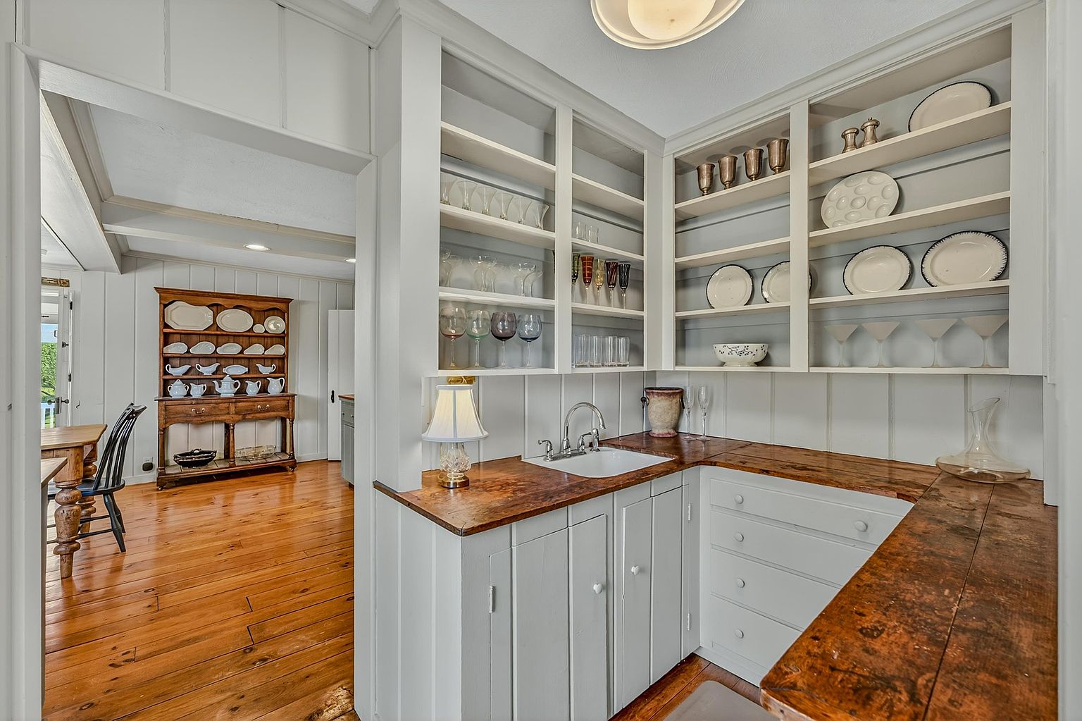 This charming butler's pantry or wet bar area features custom white cabinetry with open shelving displaying glassware and dishware. The space is anchored by a dark, rustic wood countertop and a small undermount sink, creating a warm, traditional aesthetic. Through the doorway, a glimpse of the adjacent dining area reveals a matching hutch and hardwood flooring, emphasizing the home's cohesive, classic design.