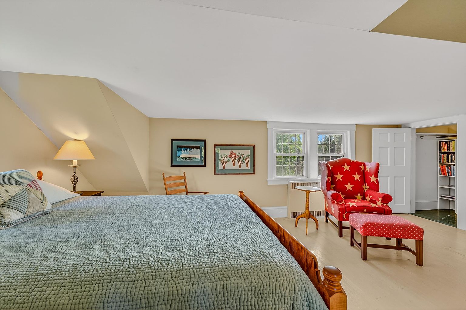 This cozy bedroom features a large bed with a textured blue quilt, positioned under a sloped ceiling characteristic of an attic space. A vibrant red armchair with star patterns and a matching ottoman create a comfortable seating area near the windows, while a small wooden chair and framed artwork add character to the room. The space is finished with light-toned flooring and neutral walls, providing a warm and inviting atmosphere.