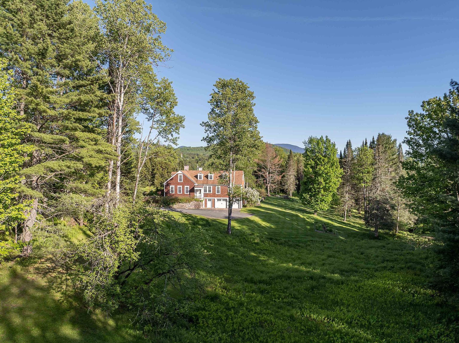 This wide-angle, elevated shot captures a traditional red-sided home nestled within a lush, expansive landscape. The property features a multi-level design with a stone foundation, a paved driveway leading to a garage, and a sprawling green lawn framed by mature trees and distant mountain vistas. The perspective emphasizes the home's secluded, picturesque setting and its integration into the natural environment.
