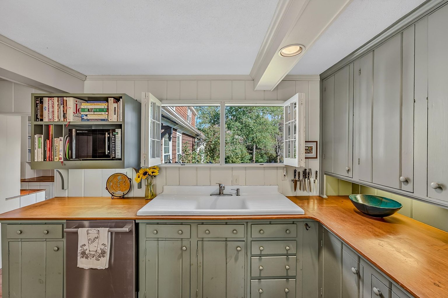 This charming kitchen features sage green cabinetry paired with warm wood countertops and a classic white farmhouse-style sink. A central window with open casements provides a view of the outdoors, while open shelving above the counter displays a collection of cookbooks. The space has a cozy, rustic aesthetic with a functional layout that includes a magnetic knife strip and integrated storage.