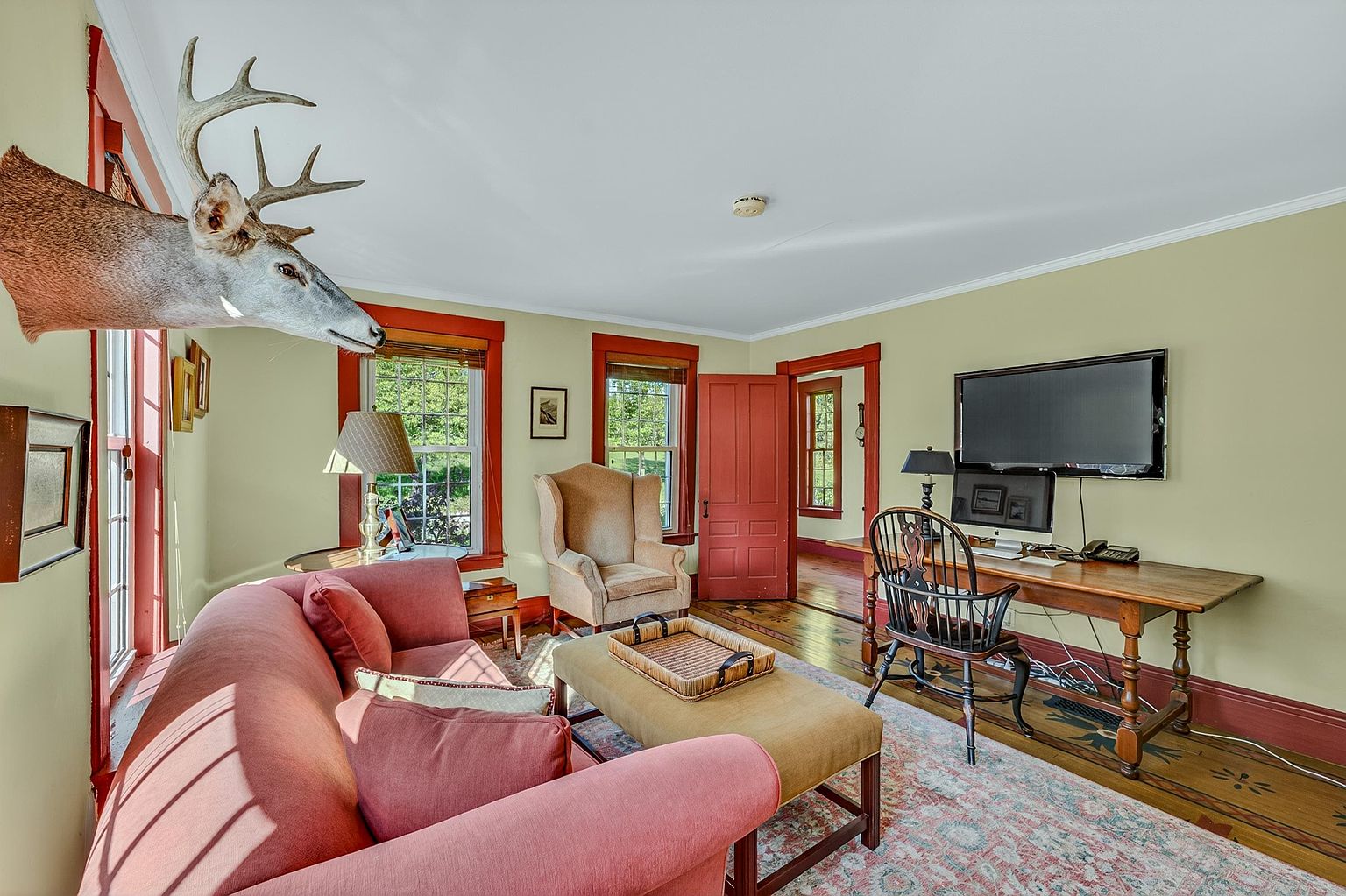 This cozy living room features a warm, traditional aesthetic with light green walls and bold red trim around the windows and doors. The space is furnished with a plush red sofa, a tan armchair, and a central ottoman, all set upon a patterned area rug over hardwood floors. A desk area with a computer monitor is positioned against the wall, while a mounted deer head serves as a prominent decorative accent on the left wall.
