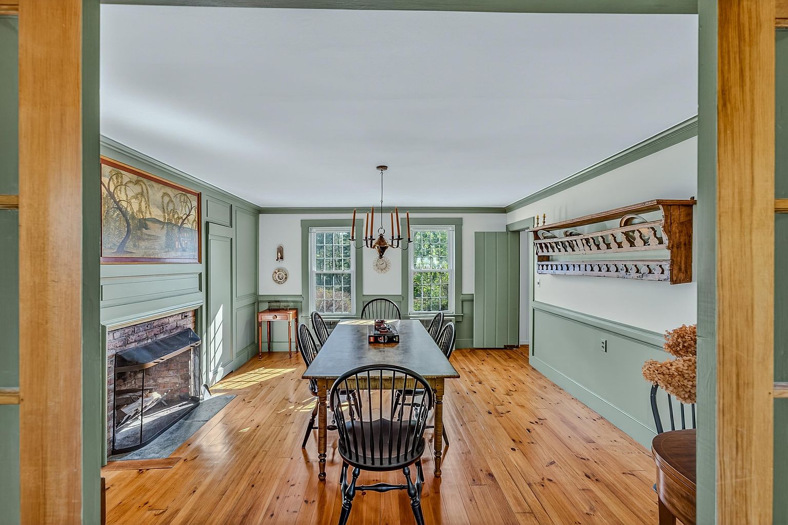This formal dining room features a rustic aesthetic with wide-plank wood flooring and sage green wainscoting that wraps around the perimeter. A long wooden table sits at the center under a traditional chandelier, flanked by a brick fireplace on the left and a decorative wooden wall-mounted shelf on the right. The perspective is framed through a doorway, looking into the space which is illuminated by natural light from two symmetrical windows.