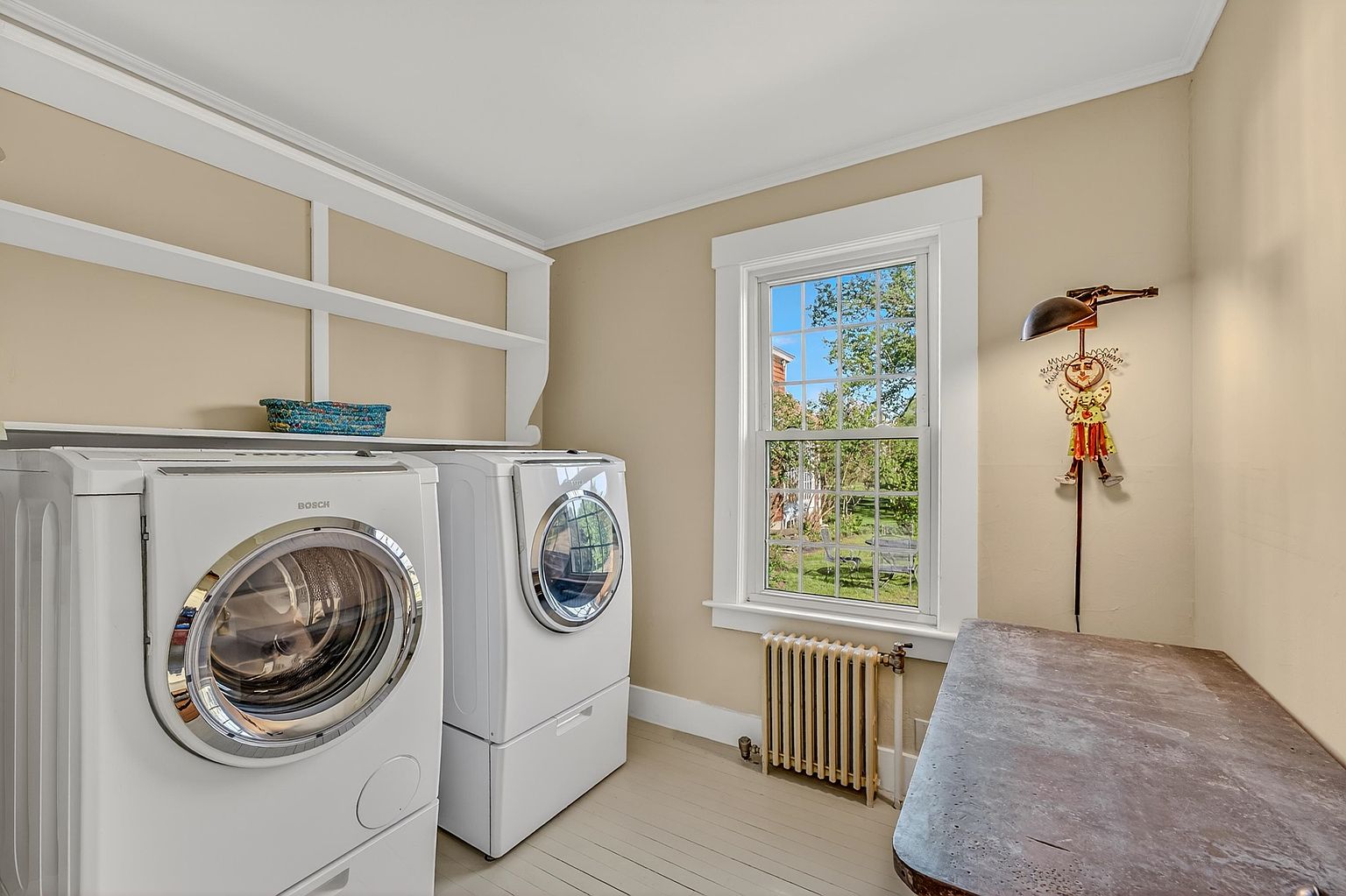 This functional laundry room features a pair of white Bosch front-loading appliances positioned beneath a built-in white shelving unit. To the right, a weathered wooden table provides a workspace, complemented by a whimsical clown-themed wall lamp. A classic radiator sits beneath a window that offers a view of the outdoor greenery, creating a bright and practical space.