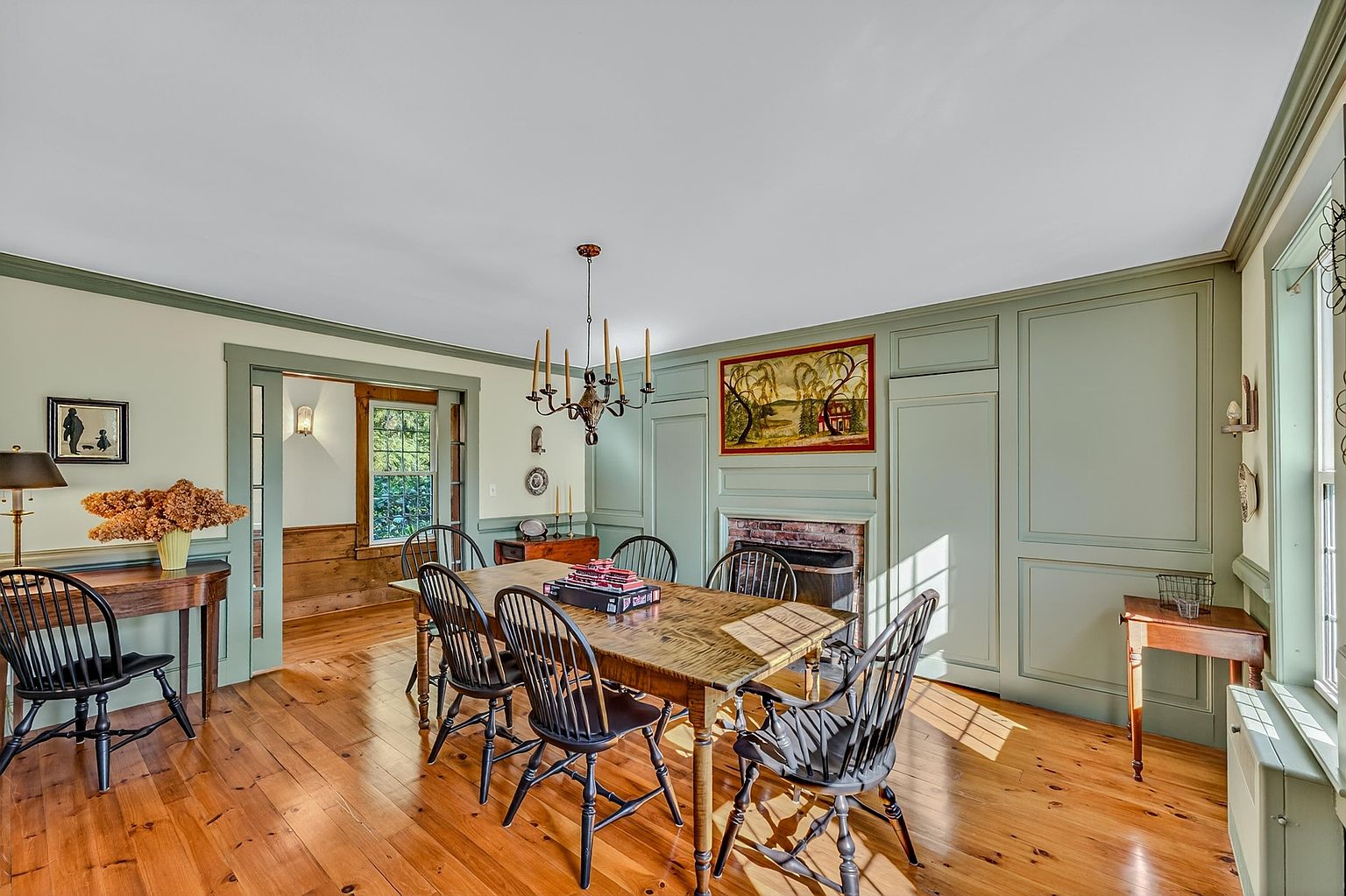 This traditional dining room features warm wood flooring and sage green paneled walls that create a cozy, historic atmosphere. A large wooden dining table is centered under a classic chandelier, surrounded by black spindle-back chairs. A brick fireplace surround adds a rustic focal point, while an adjacent doorway leads into a sunlit room with natural wood wainscoting.