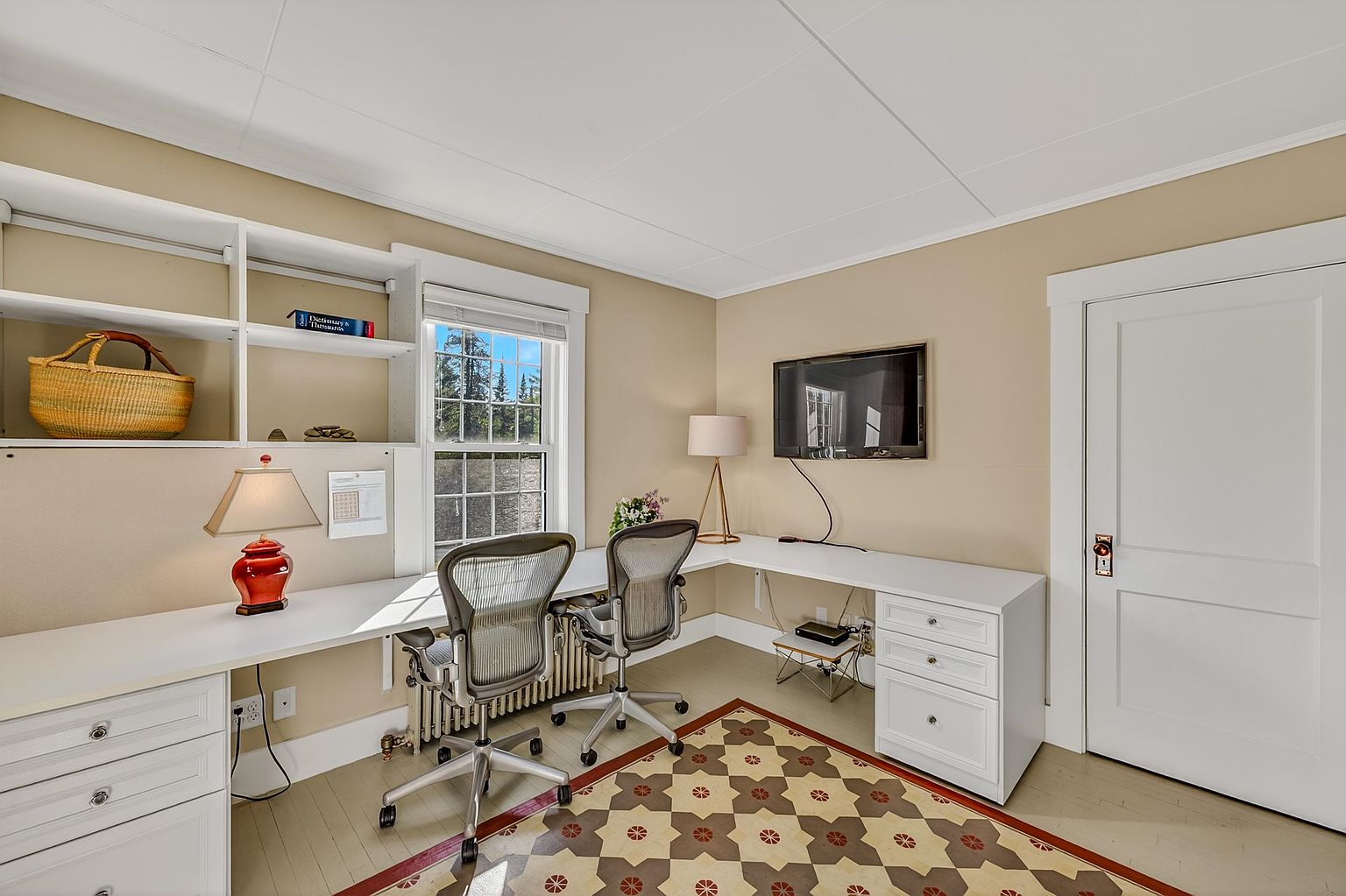 This bright and functional home office features a large L-shaped white desk with built-in storage drawers and shelving units. Two ergonomic office chairs are positioned at the desk, which sits beneath a window that allows for natural light, while a patterned area rug adds visual interest to the room. The space is completed by a wall-mounted television and a table lamp, creating a clean and organized workspace.