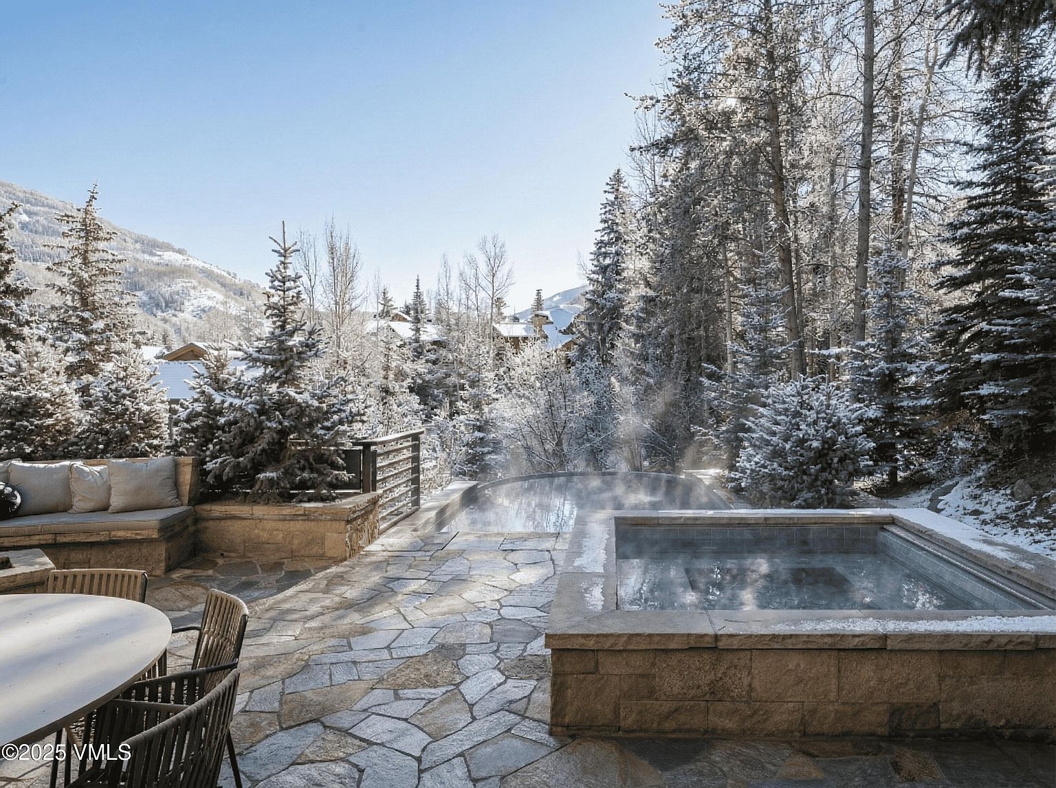 This image showcases a luxurious outdoor patio area in a snowy, mountainous setting. The patio features a stone surface, a built-in seating area with cushions, and a hot tub with steam rising from the water. The surrounding landscape includes snow-covered trees and a clear blue sky, creating a serene and inviting atmosphere.