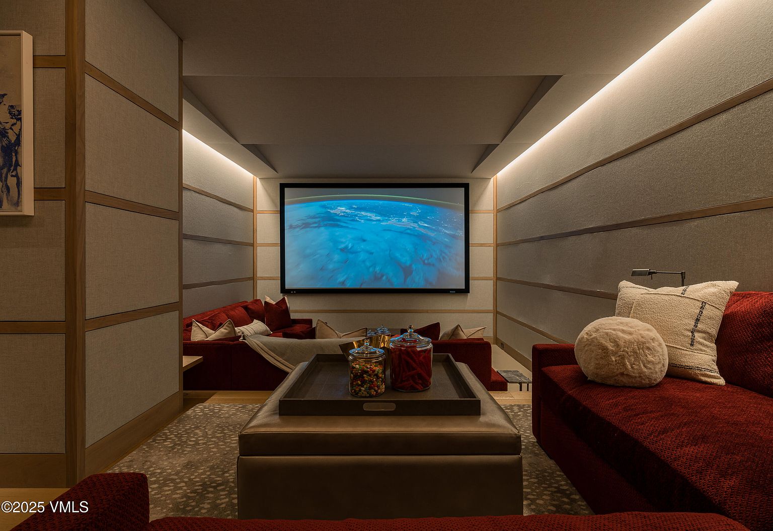 This is an interior shot of a home theater or living room, featuring a large screen displaying an image of Earth. The room is designed with sound-dampening panels and recessed lighting, creating a cozy and immersive atmosphere. Comfortable red couches and a central ottoman with candy jars invite relaxation and entertainment.
