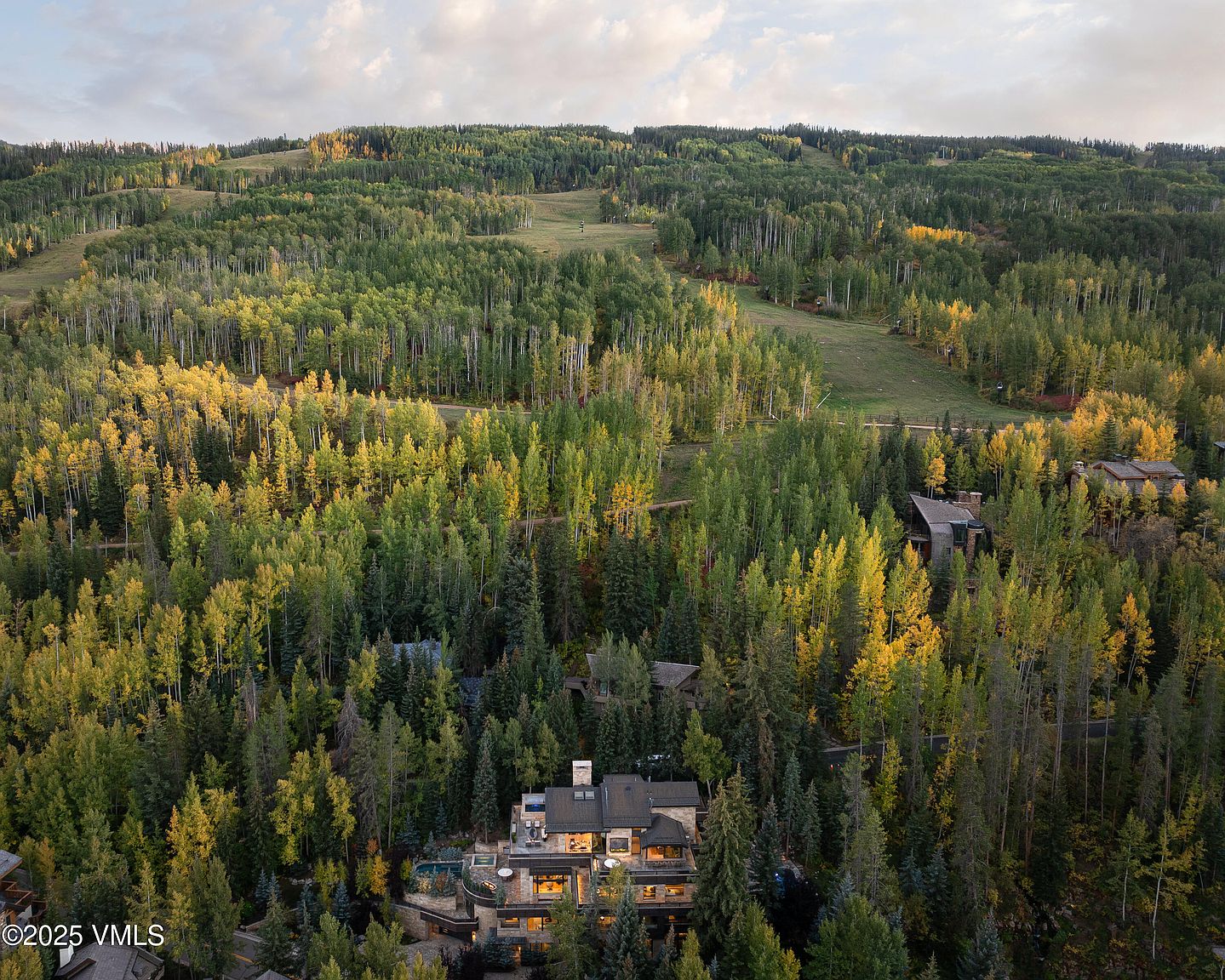 This aerial view showcases a luxurious home nestled amidst a dense forest with trees displaying vibrant fall colors. The house features a stone exterior, multiple levels, and a well-maintained landscape including a pool area. The surrounding forest extends up a hillside, creating a sense of privacy and natural beauty.