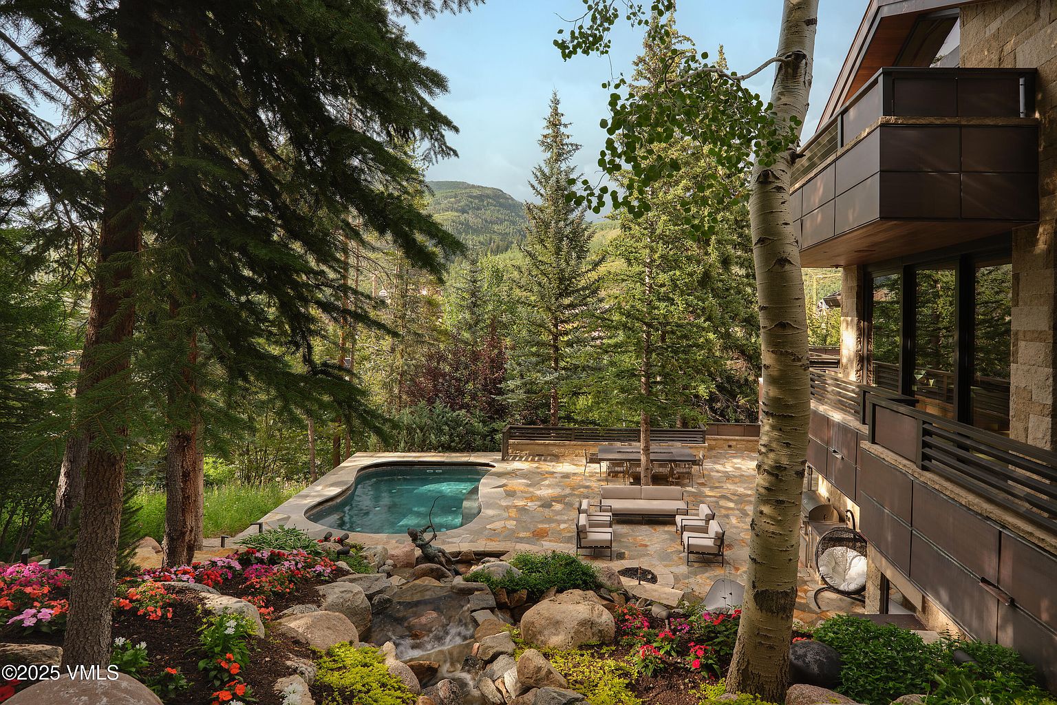 This image showcases a luxurious outdoor space featuring a kidney-shaped pool with clear blue water, surrounded by a stone patio with elegant outdoor furniture. A natural rock waterfall flows into the pool, adding to the serene ambiance, while lush landscaping with colorful flowers and mature trees provides privacy and enhances the property's appeal. The adjacent modern home with balconies complements the outdoor living area, creating an inviting and upscale retreat.