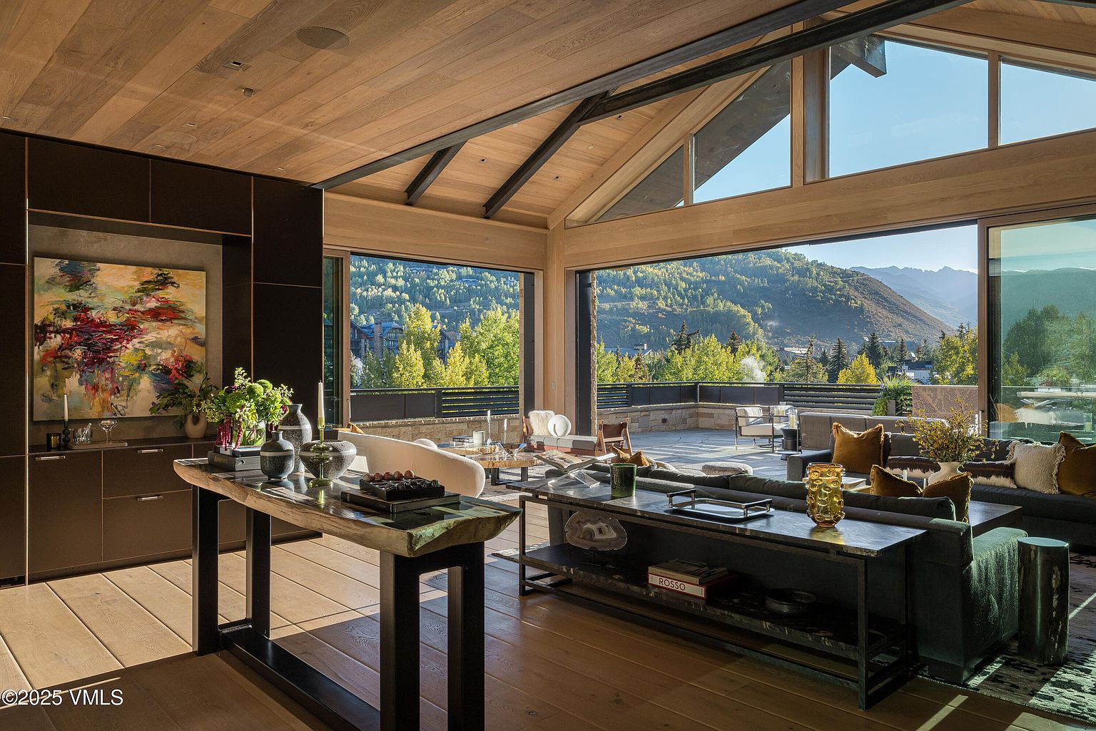 This is a spacious living room with a modern design, featuring a high, wood-paneled ceiling with exposed beams and large sliding glass doors that open to a patio with mountain views. The room is furnished with a comfortable gray sofa, decorative pillows, and stylish coffee tables, creating an inviting and luxurious atmosphere. An abstract painting and decorative objects add to the room's sophisticated aesthetic.
