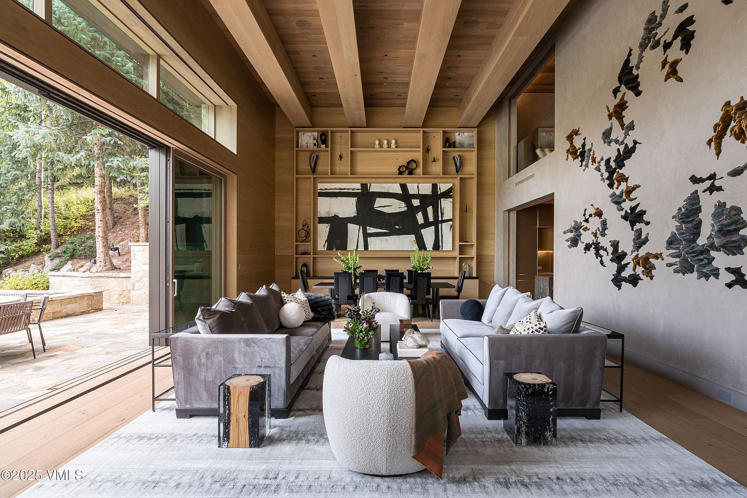 This is an interior shot of a luxurious living room featuring two gray sofas, a unique textured chair, and striking artwork. The room boasts a high ceiling with wooden beams and a large open doorway leading to an outdoor patio. The overall impression is one of modern elegance and spaciousness, perfect for entertaining or relaxing.