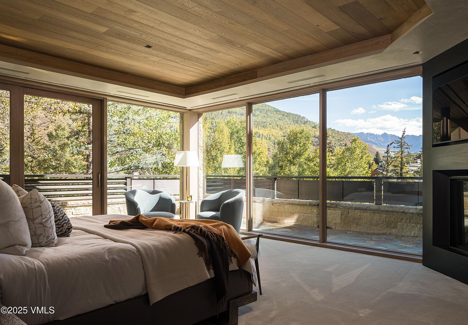 This is a luxurious primary bedroom featuring a king-size bed with plush bedding and a throw blanket. The room boasts floor-to-ceiling windows offering stunning views of the surrounding mountains and trees. Two armchairs and a side table are positioned near the windows, creating a cozy seating area. The ceiling is finished with wood panels, adding warmth and texture to the space.