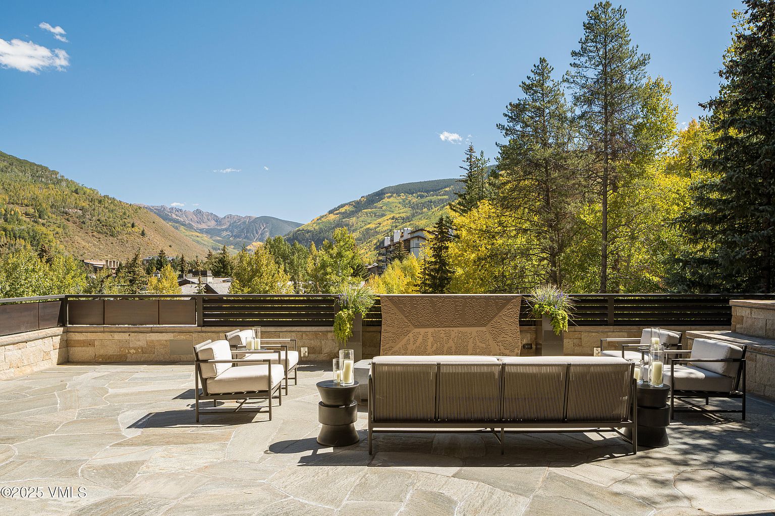 This image showcases a luxurious patio or deck area with comfortable outdoor seating, including armchairs and a sofa, arranged around small side tables. The patio is paved with large stone tiles, and a low wall with a decorative screen provides a sense of enclosure while still offering views of the surrounding mountains and trees. The overall impression is one of sophisticated outdoor living and relaxation.