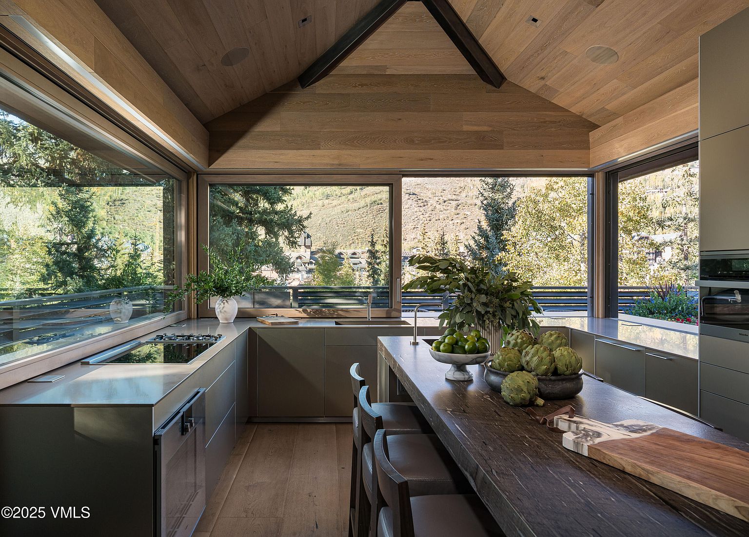 This interior shot showcases a modern kitchen with a rustic touch, featuring sleek gray cabinetry and a dark wood island with bar seating. Large windows offer stunning views of the surrounding landscape, bringing natural light into the space. The kitchen's design blends contemporary elements with natural materials, creating a warm and inviting atmosphere.