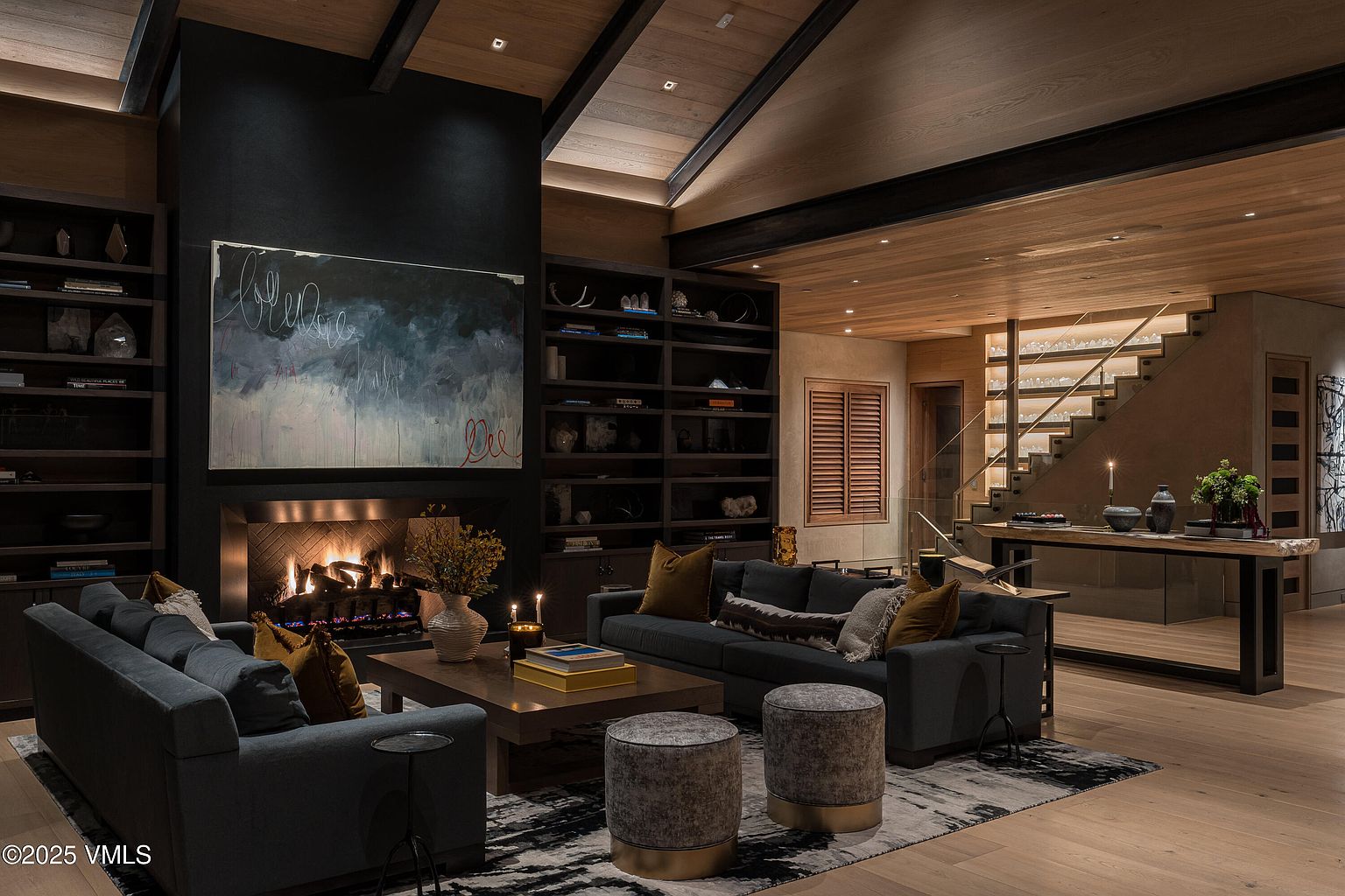 This is an interior shot of a luxurious living room featuring a stone fireplace with a large abstract painting above it. The room is furnished with two dark-colored sofas, a wooden coffee table, and two upholstered ottomans on a patterned rug. The space is well-lit, highlighting the wooden ceiling beams and the staircase in the background, creating a warm and inviting atmosphere.