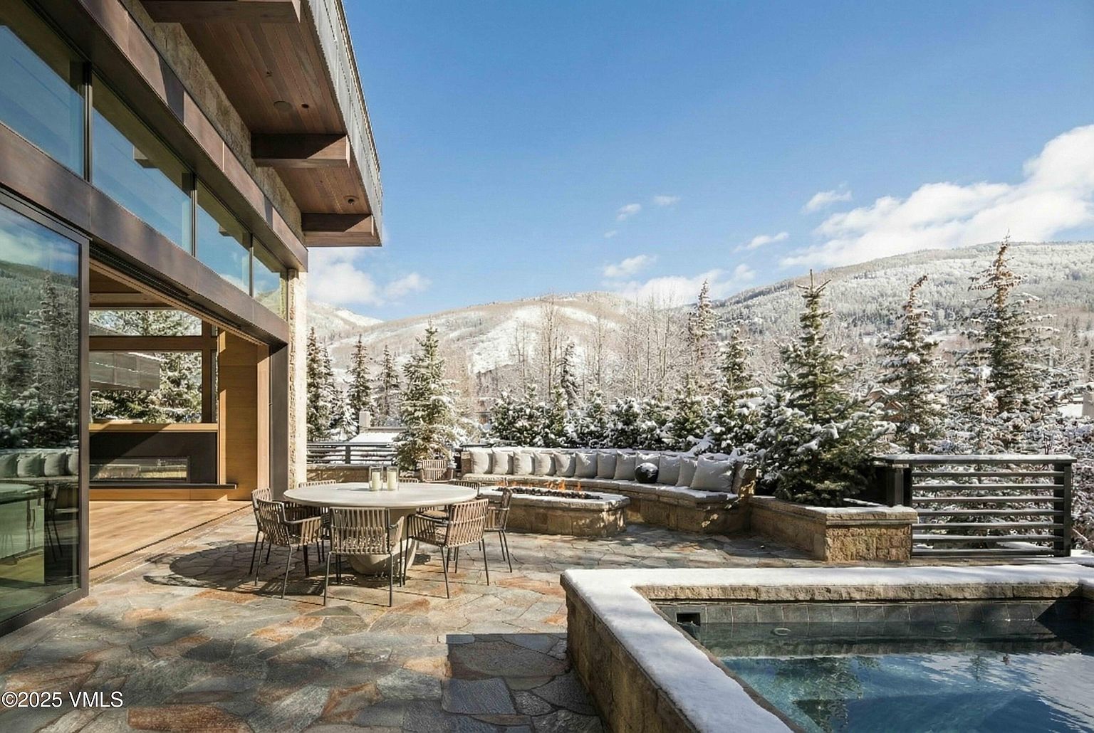 This image showcases a luxurious outdoor patio area with a stone-paved surface, a round dining table with chairs, and a built-in seating area with a fire pit. A pool or spa is visible in the foreground, and snow-covered trees and mountains provide a scenic backdrop. The patio is adjacent to a modern home with large glass windows, creating a seamless indoor-outdoor living experience.