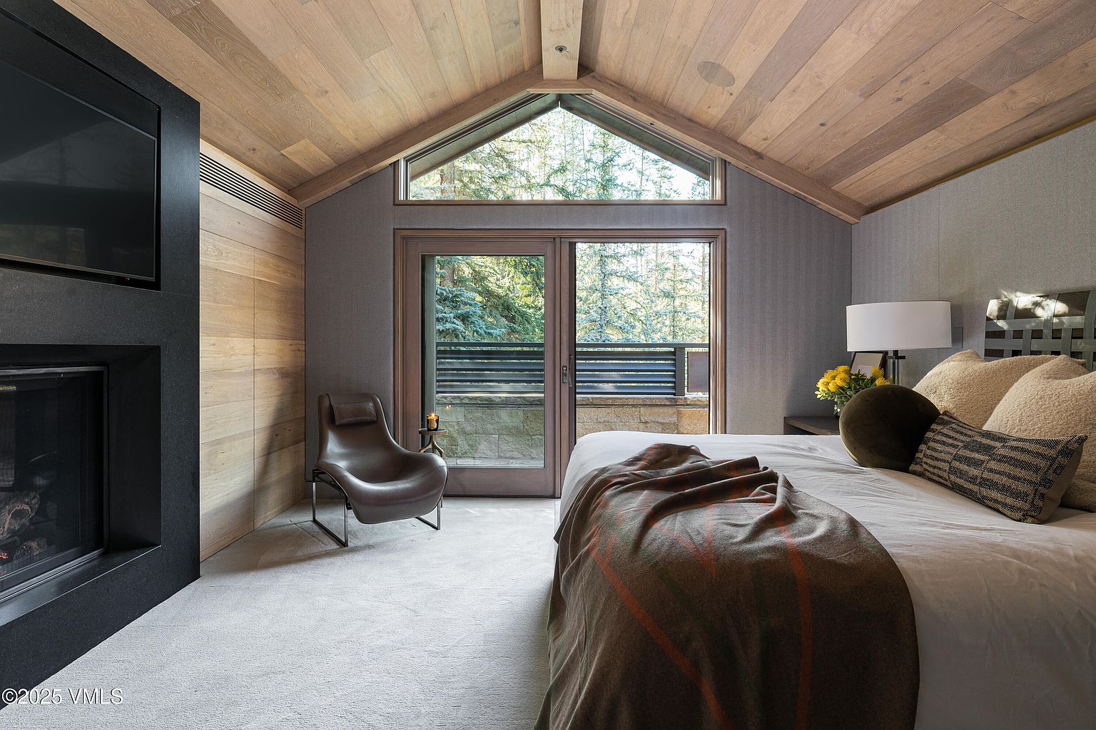 This is a primary bedroom featuring a modern design with a fireplace and a large window overlooking a wooded area. The room has a neutral color palette with wood accents on the ceiling and walls, creating a warm and inviting atmosphere. A comfortable chair sits near the window, and the bed is neatly made with decorative pillows and a throw blanket.