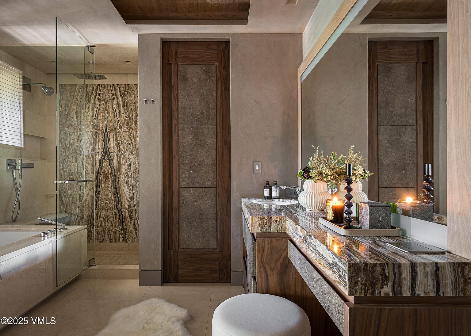 This is a luxurious primary bathroom featuring a marble countertop vanity with a large mirror, a modern sink, and decorative vases. A glass-enclosed shower with marble walls adds to the spa-like atmosphere. The room is well-lit and exudes a sense of elegance and relaxation.