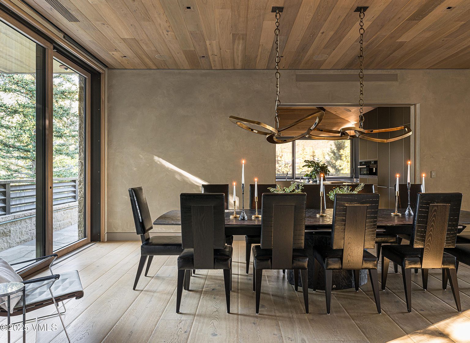 This is an interior shot of a dining room featuring a long, dark wood table surrounded by black upholstered chairs. A modern chandelier hangs above the table, which is adorned with candles and greenery. A large sliding glass door provides natural light and a view to the outside, while the wood plank ceiling adds warmth to the space.