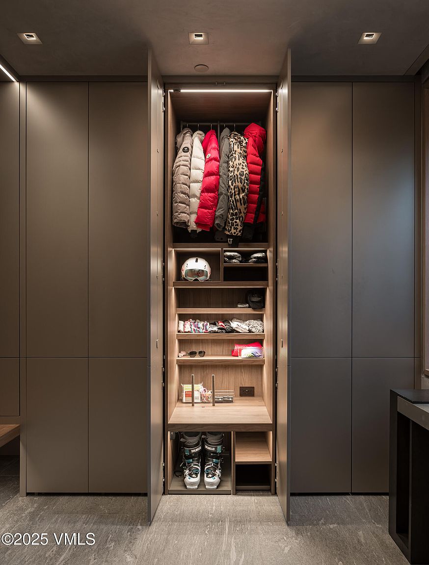 This image showcases a well-organized walk-in closet with a modern design. The closet features custom shelving and hanging rods, filled with neatly arranged clothing, ski gear, and accessories. Integrated lighting enhances the space, creating a clean and functional storage solution.