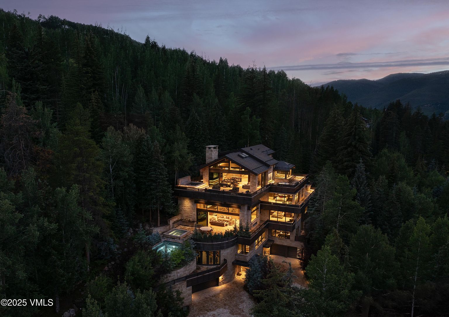 This aerial view showcases a luxurious multi-story home nestled in a dense forest at dusk. The house features multiple balconies, large windows revealing warmly lit interiors, and a pool/spa area. The architectural style is modern and sophisticated, blending seamlessly with the natural surroundings, creating an impression of secluded elegance and high-end living.