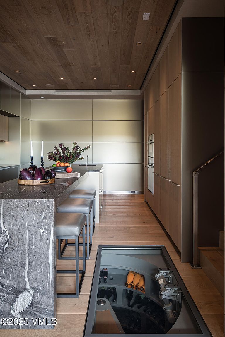 This is a modern kitchen featuring sleek, minimalist cabinetry and a striking marble island with bar seating. A unique glass-covered wine cellar is integrated into the floor, adding a luxurious touch. The warm wood flooring and ceiling create a sophisticated and inviting atmosphere.
