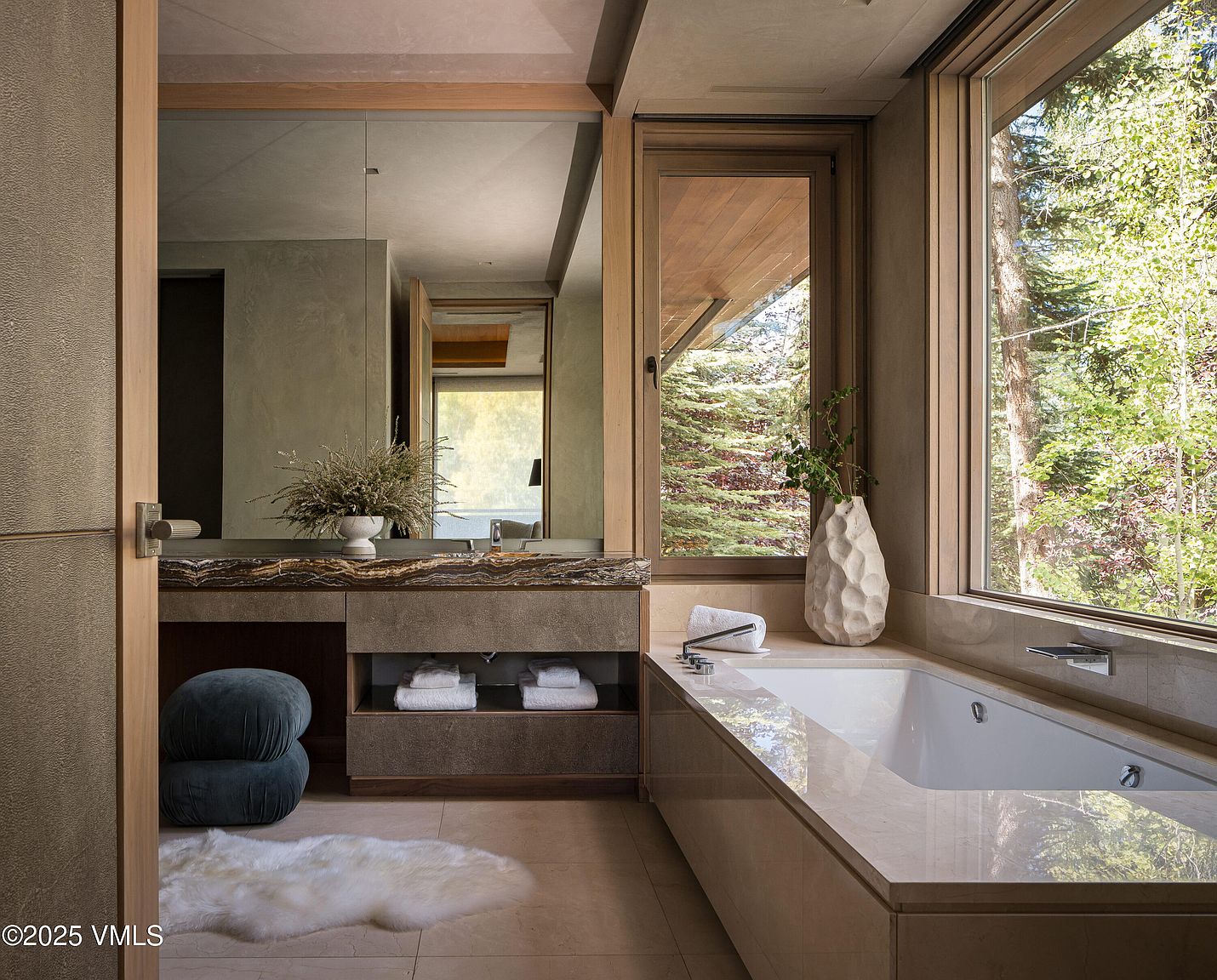 This is a luxurious primary bathroom featuring a large soaking tub positioned next to a window with a view of lush greenery. The vanity area includes a marble countertop, a large mirror, and built-in shelving for towels. The overall design is modern and serene, creating a spa-like atmosphere.