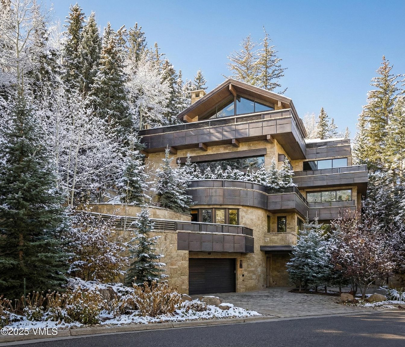This is a front exterior view of a modern mountain home. The house features a stone facade, multiple balconies with metal railings, and large windows. The surrounding landscape includes snow-covered trees, adding to the property's appeal and highlighting its location in a winter setting.