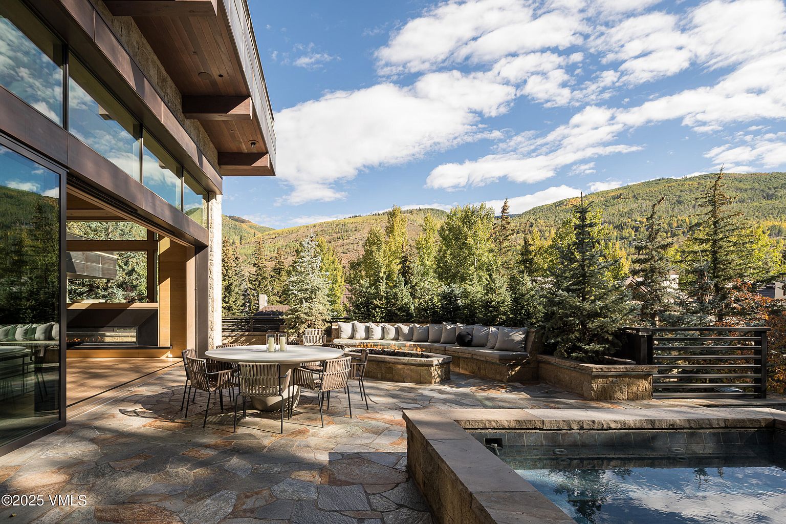 This image showcases a luxurious outdoor patio area, featuring a stone-paved surface, a round dining table with chairs, and a built-in seating area with a fire pit. A swimming pool is partially visible in the foreground, and the backdrop includes lush greenery and mountains, creating a serene and upscale ambiance. The open glass doors lead into the house, blending indoor and outdoor living.