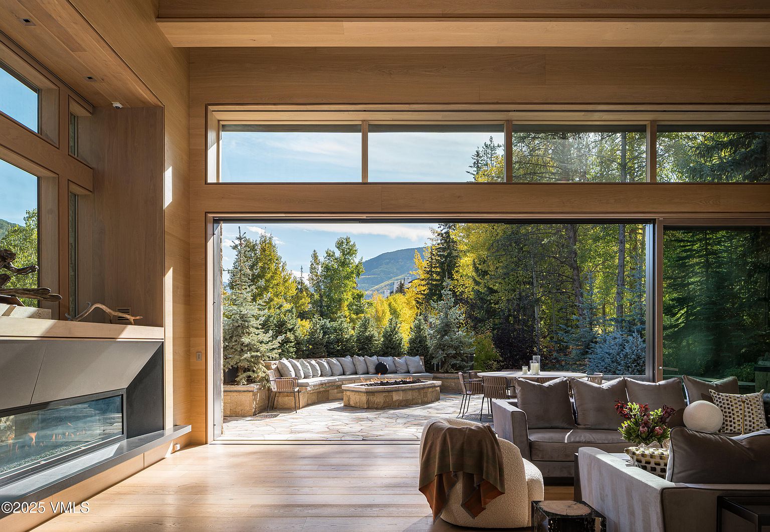 This is a luxurious living room that seamlessly blends indoor and outdoor living. The room features a modern fireplace, hardwood floors, and large sliding glass doors that open to a patio with a fire pit and seating area. The interior is furnished with comfortable sofas and chairs, creating an inviting and sophisticated space.