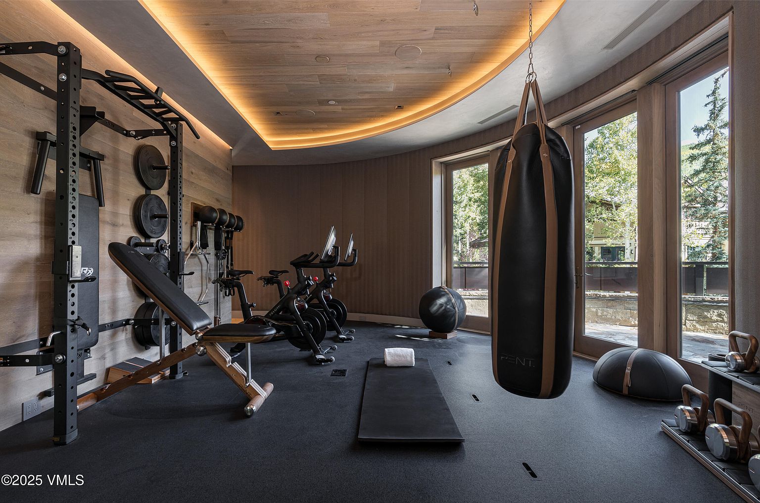 This is a well-equipped home gym featuring a weight rack, adjustable bench, stationary bikes, a punching bag, and various exercise balls and kettlebells. The room has a modern aesthetic with wood paneling, recessed lighting, and large windows offering natural light and views of the outdoors. The black rubber flooring provides a functional and safe workout space.