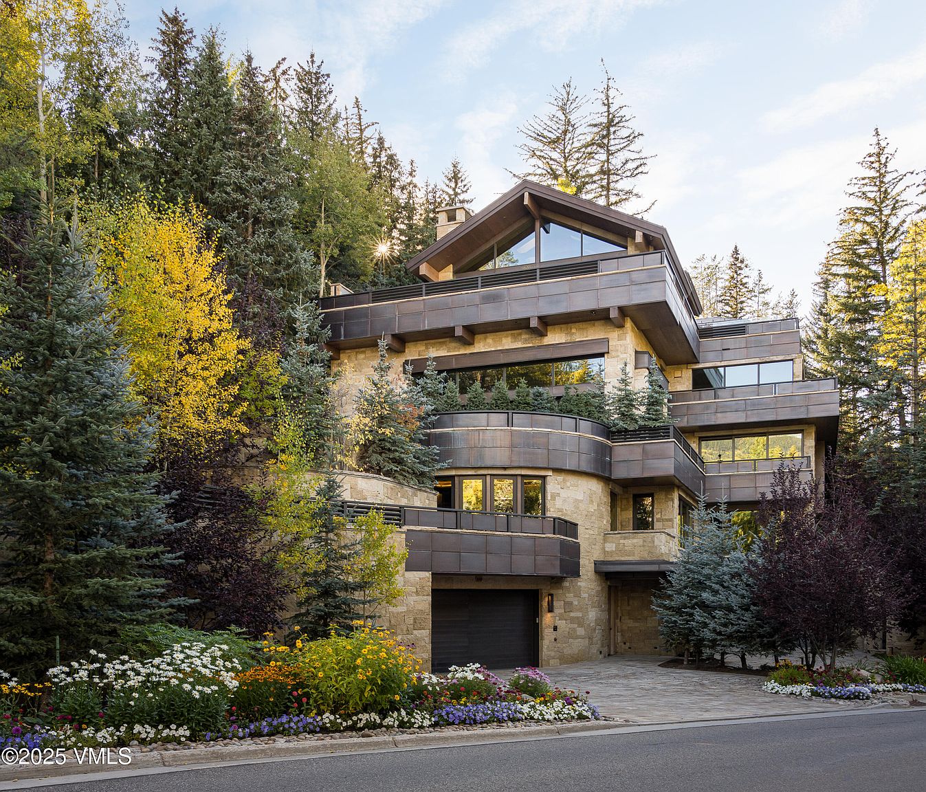 This is a front exterior view of a modern, multi-level home nestled among lush trees and landscaping. The house features a combination of stone and metal exterior finishes, with large windows and balconies. A well-maintained driveway leads to a garage, and colorful flowerbeds add to the curb appeal.