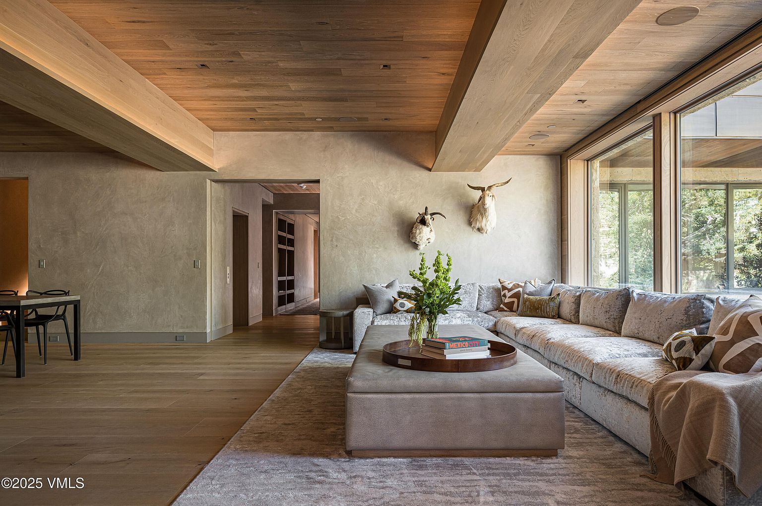 This is an interior shot of a luxurious living room featuring a large, comfortable sectional sofa with decorative pillows, a plush ottoman serving as a coffee table, and a neutral-toned rug. The room is adorned with unique wall decor, including mounted animal heads, and boasts a wooden ceiling with exposed beams. Large windows provide ample natural light and a view of the outdoors, creating a warm and inviting atmosphere.