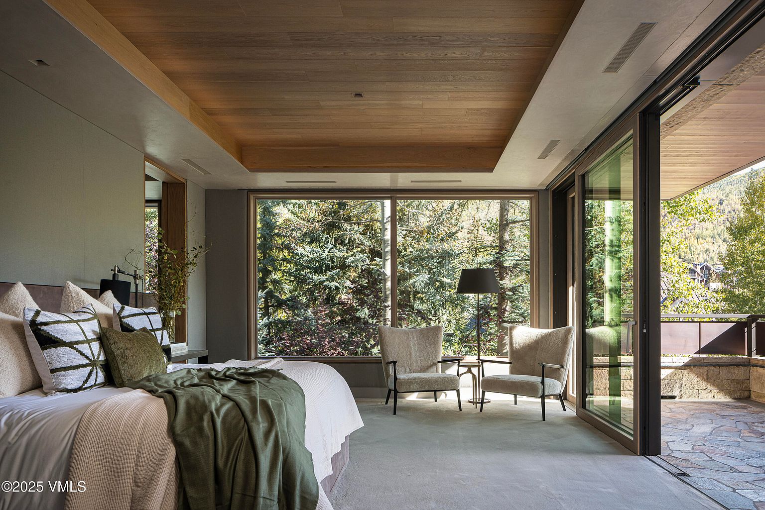 This is a luxurious primary bedroom featuring a large bed with plush pillows and a throw blanket. The room has a modern design with a wooden ceiling and large windows offering a view of the surrounding forest. A pair of armchairs and a small table create a cozy seating area near the windows, and sliding glass doors lead to an outdoor patio.