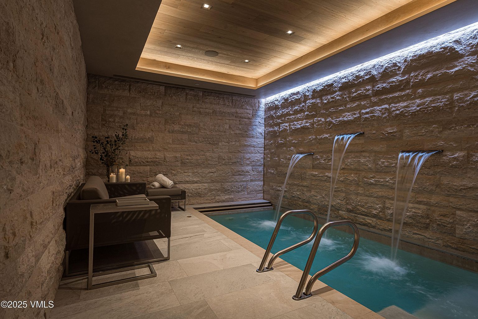 This image showcases a luxurious indoor spa area featuring a small pool with three waterfall features cascading from a textured stone wall. The space includes a seating area with a modern sofa and side table, complemented by soft lighting from the wooden ceiling and recessed lights. The overall impression is one of tranquility and high-end relaxation.