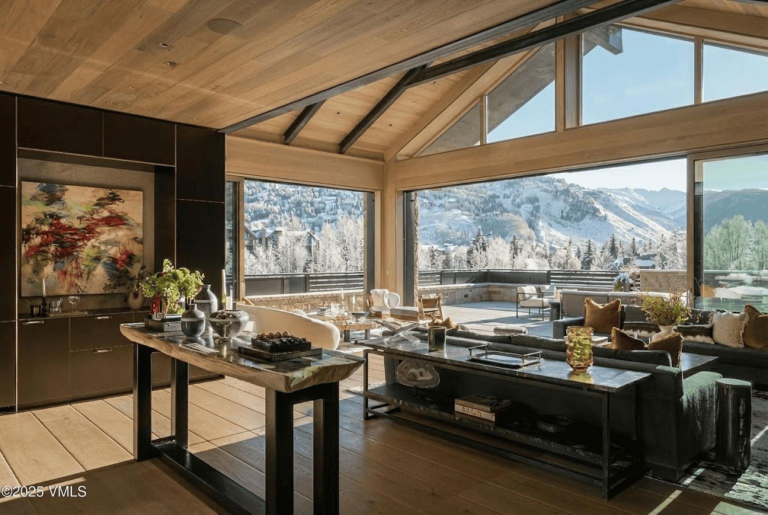 This is an interior shot of a luxurious living room featuring a high vaulted ceiling with wooden beams and large windows offering a stunning view of snow-covered mountains. The room is furnished with modern sofas, stylish tables, and decorative items, creating a sophisticated and inviting atmosphere. The open layout seamlessly connects the indoor space with an outdoor patio, enhancing the sense of spaciousness and natural light.
