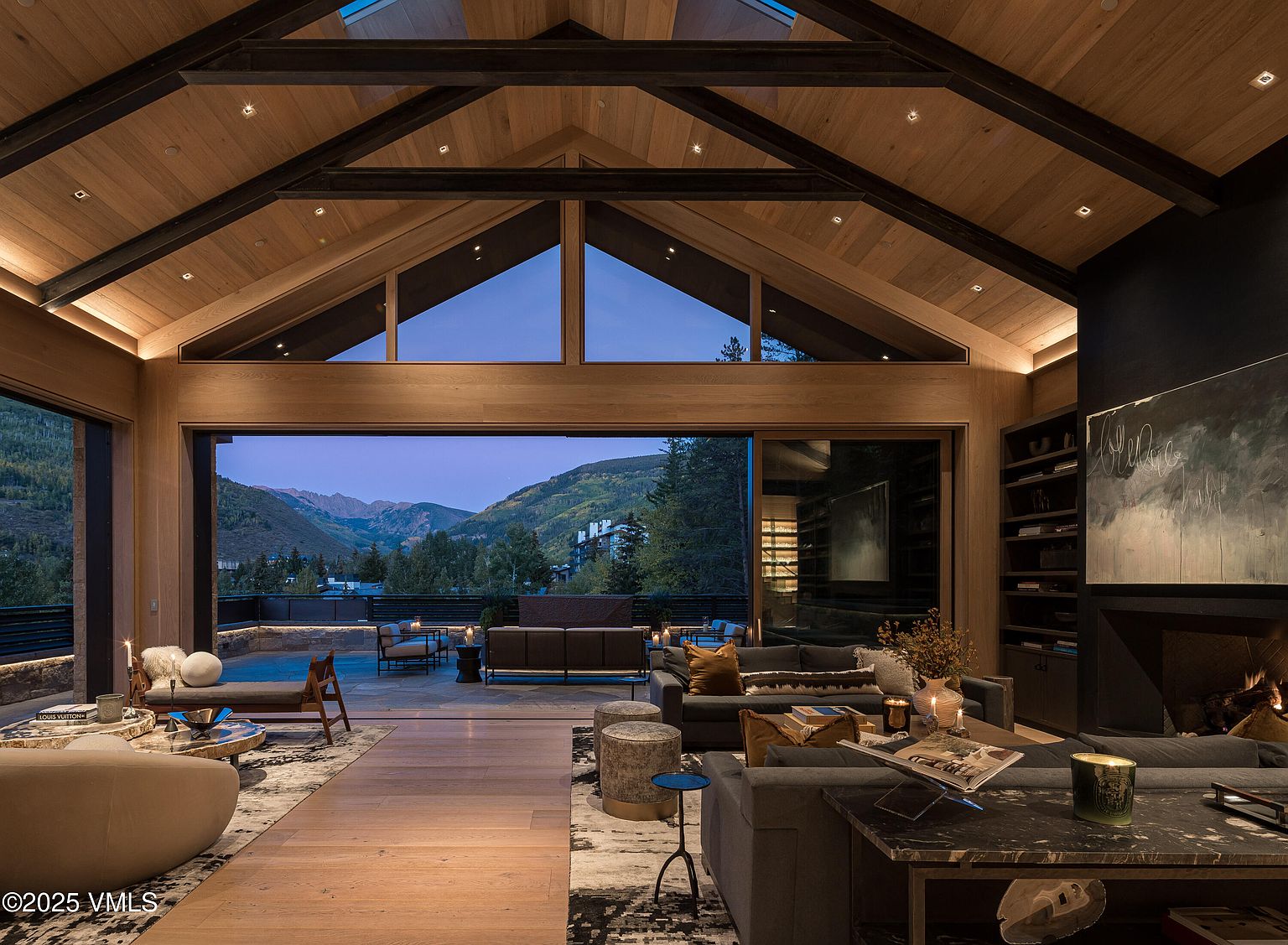 This is a luxurious living room featuring a high vaulted ceiling with wooden beams and paneling, creating a warm and inviting atmosphere. Large sliding glass doors open to a patio with mountain views, seamlessly blending indoor and outdoor living. The room is furnished with comfortable sofas, stylish coffee tables, and a fireplace, offering a perfect space for relaxation and entertainment.