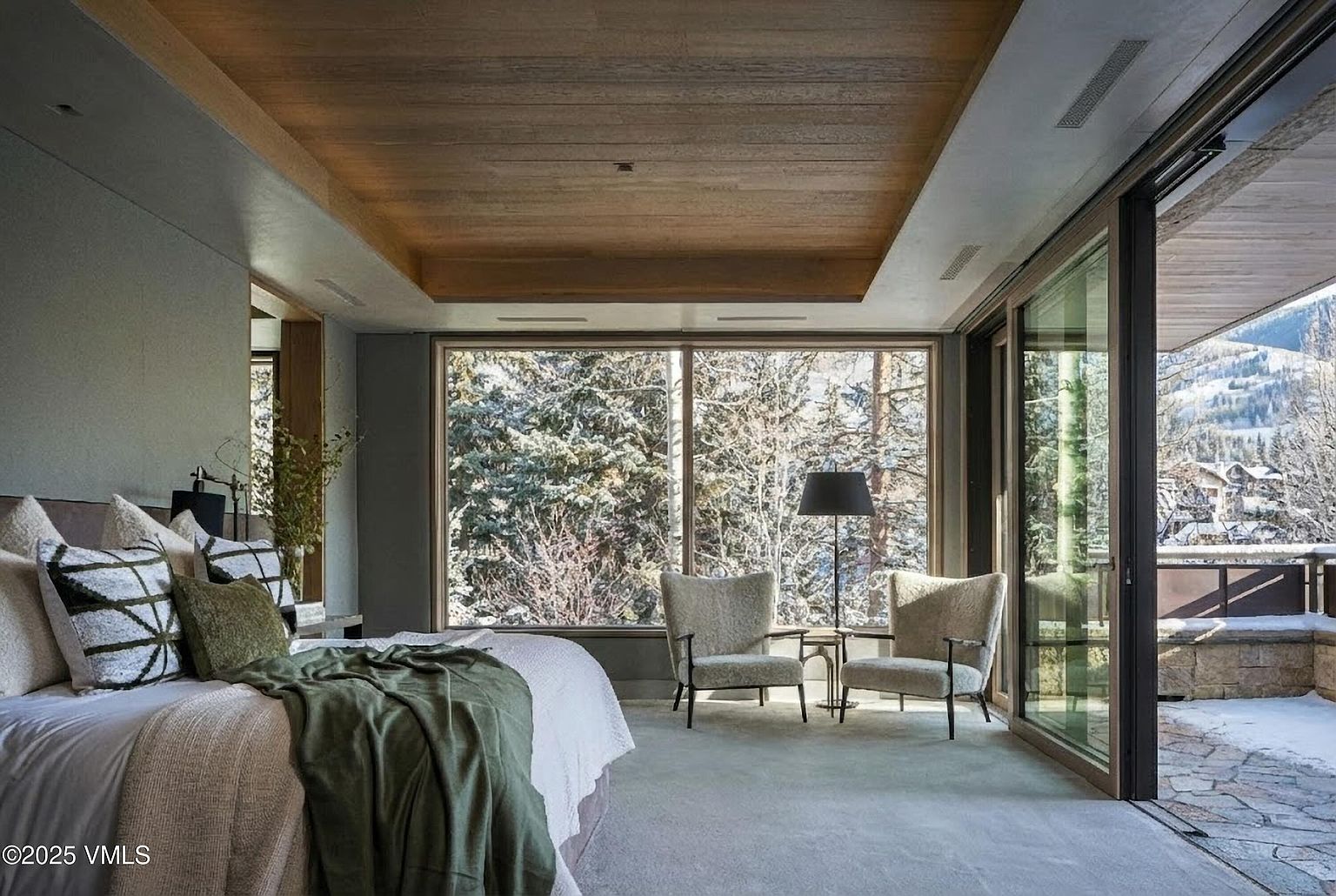 This primary bedroom features a serene and luxurious design with a focus on natural light and views. A large window frames a snowy forest scene, complemented by a seating area with two armchairs. The bed is adorned with plush pillows and a throw, adding to the room's cozy and inviting atmosphere, while a sliding glass door leads to an outdoor patio.