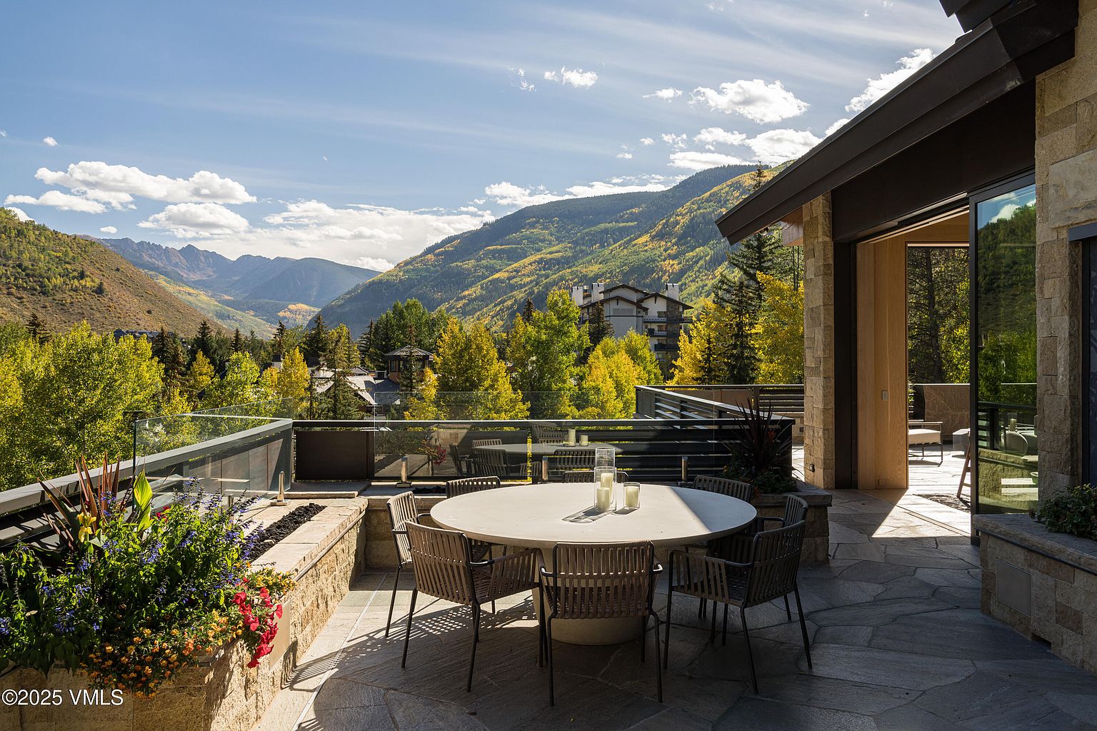This image showcases a luxurious outdoor patio or deck area with stunning mountain views. A round dining table with chairs is centrally located, suggesting an ideal space for outdoor dining and entertaining. The patio features a stone floor and a built-in planter with vibrant flowers, adding to the upscale ambiance.