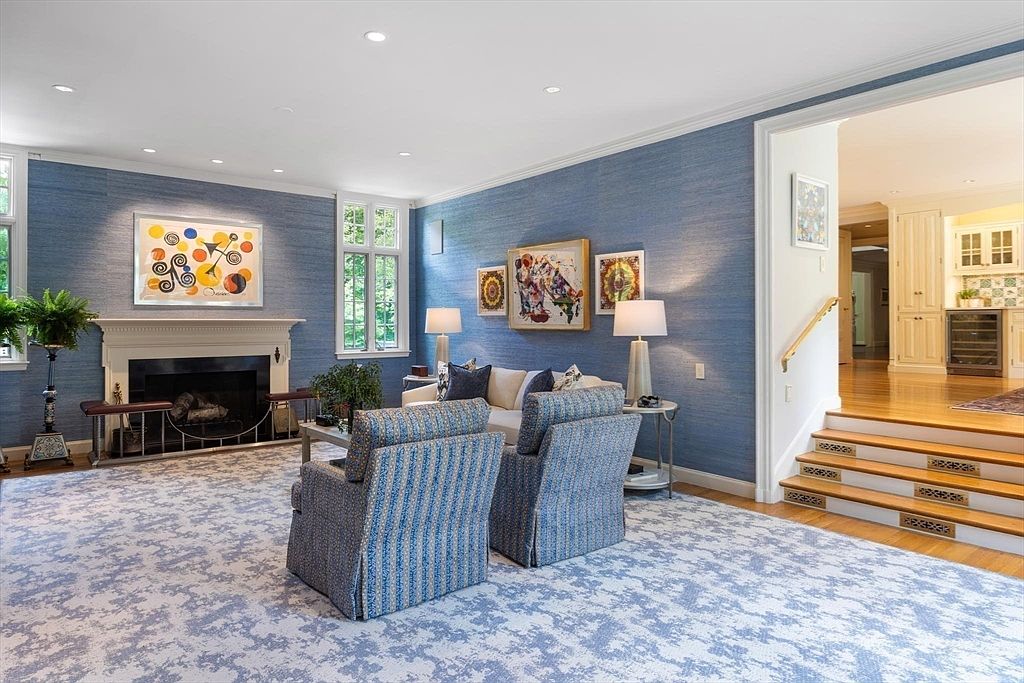 This elegant living room features textured blue wallpaper, a classic fireplace with a decorative mantel, and a plush patterned carpet. The space is furnished with two comfortable armchairs and a sofa, accented by abstract artwork and warm table lamps. A set of wooden stairs leads up to an adjacent room, creating an inviting and sophisticated atmosphere.