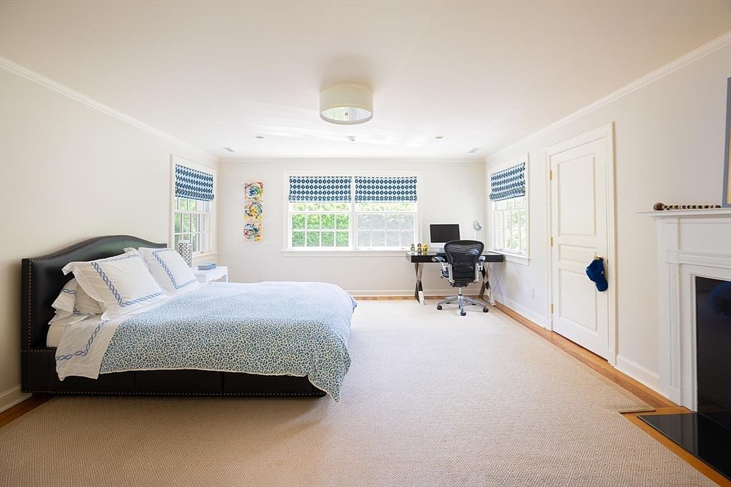 This spacious primary bedroom features a large bed with a dark upholstered headboard and a blue patterned duvet, set against a neutral color palette. The room includes a dedicated workspace with a desk and ergonomic chair near the windows, as well as a fireplace on the right side. The wide-angle perspective captures the airy, well-lit atmosphere, highlighting the blend of comfort and functionality.