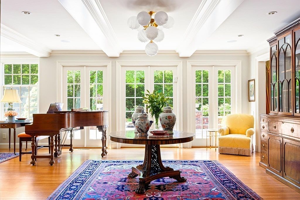 This elegant and spacious living room features a grand piano, a central round wooden table adorned with decorative vases, and a vibrant, traditional area rug. The room is bathed in natural light from multiple sets of French doors that open to a lush garden view, complemented by a sophisticated chandelier and a classic yellow armchair. The space exudes a refined, traditional aesthetic, perfect for formal entertaining or quiet relaxation.