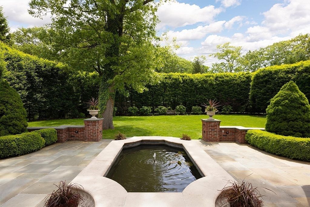 This elegant backyard features a stone-paved patio centered around a formal, octagonal water fountain with a gentle spray. The manicured lawn is framed by tall, dense privacy hedges and mature trees, creating a serene and secluded atmosphere. The scene is captured from a low, eye-level perspective, emphasizing the symmetry and sophisticated landscaping of the outdoor space.