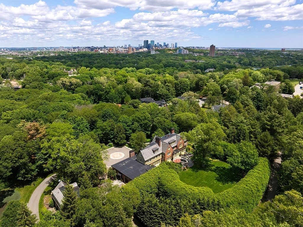 This high-angle aerial view captures a grand, multi-story brick estate nestled within a lush, dense forest canopy. The property features a circular driveway, a well-manicured lawn, and a sprawling backyard, all set against the distant, panoramic skyline of a major city. The perspective emphasizes the home's secluded, private setting while highlighting its proximity to urban amenities.