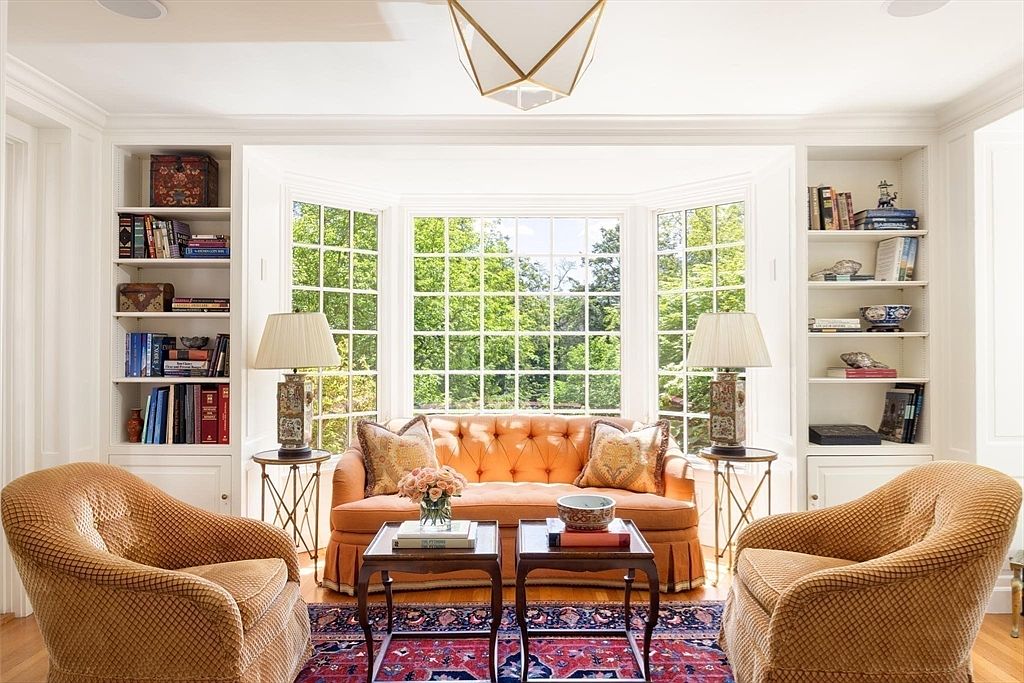 This elegant living room features a symmetrical layout centered around a vibrant orange tufted sofa positioned in front of a large, light-filled bay window. Flanking the window are built-in white bookshelves filled with books and decorative objects, while two textured armchairs and a patterned area rug complete the cozy, traditional seating area. The space is illuminated by a geometric pendant light and two matching table lamps, creating a warm and inviting atmosphere.